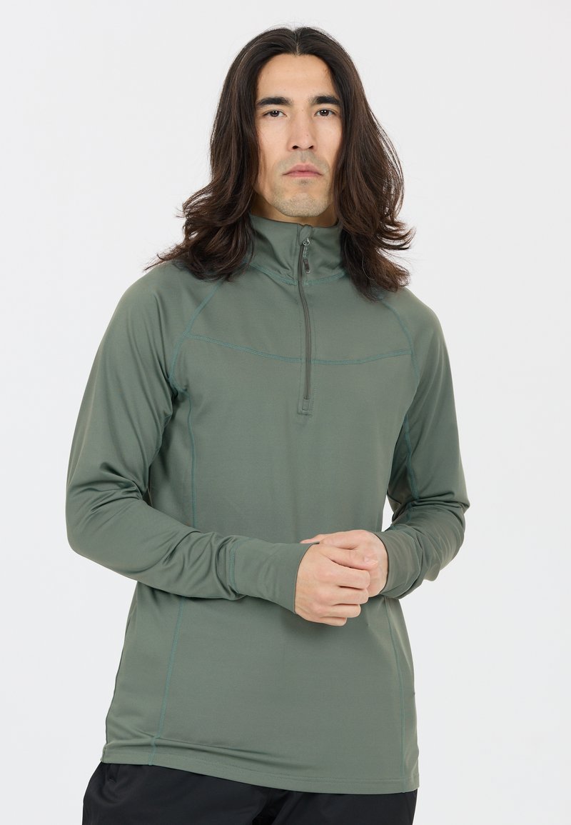 Mann mit langem dunklem Haar, der einen olivgrünen Pullover mit Reißverschluss und schwarze Hosen trägt, vor einem schlichten weißen Hintergrund stehend.