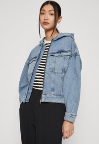 Denim striukė su kapišonu, šviesiai mėlynos spalvos, su priekinėmis kišenėmis ir užtrauktuku. Dėvima ant juodai baltos juostuotos marškinėlių.