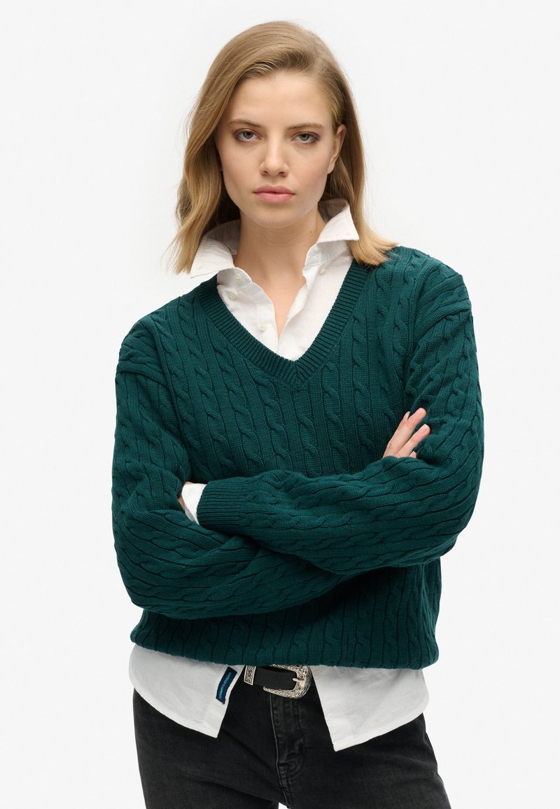 Pull en maille torsadée teal avec un col en V, porté sur une chemise blanche à col. Il a des poignets et un ourlet côtelés, associé à un jeans foncé et une ceinture en argent.