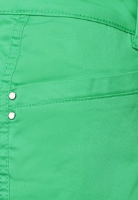 Grön tyg med en slät textur och lätt lyster. Har synlig sömnad och två metallaccenter nära fickan.