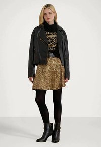 Jaqueta de couro preta sobre um suéter preto com padrão, combinada com uma saia dourada cheia de lantejoulas e collants pretos. Botas altas pretas completam o visual.