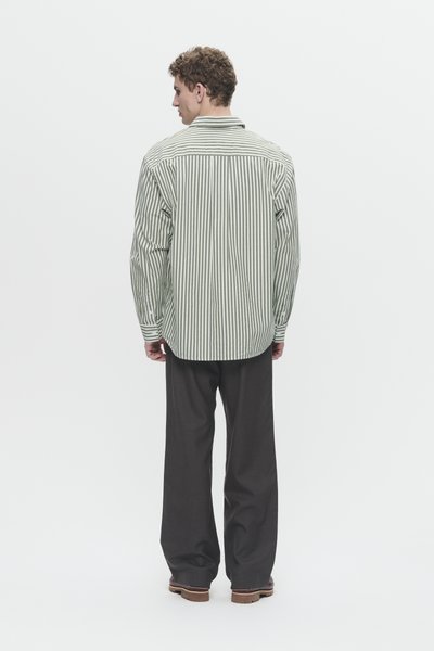Uomo di spalle che indossa una camicia a righe verticali verdi e bianche, pantaloni larghi neri e scarpe marroni su uno sfondo bianco.