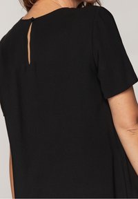 Femme portant une robe noire à manches courtes avec une petite ouverture en forme de goutte et une fermeture par bouton à l'arrière du col.