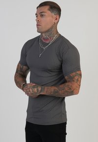Hallide ribidega lühikeste varrukatega hall särk, mis on keha kallale sobiv. Model on näidanud tätoveeritud käsi ja kihilisi hõbedaseid kaelaketid. Seljas on mustad püksid.