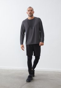 Donker langemouwen sportshirt met een mesh paneel, gecombineerd met zwarte shorts en aansluitende zwarte leggings. Model draagt zwarte sportschoenen.