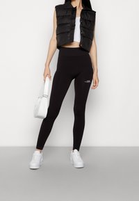 Schwarze Pufferweste über einem weißen Crop-Top, kombiniert mit hochgeschnittenen schwarzen Leggings mit einem Logo. Weiße Sneaker und eine kleine weiße Handtasche.
