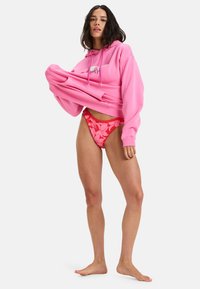 Rosa oversized hoodie med dragsko, matchad med röda bikiniunderdelar med blommigt mönster. Modellen står barfota och håller i hoodien.