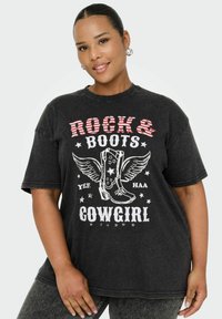 Uśmiechnięta kobieta w czarnej koszulce z napisem "Rock & Boots Cowgirl" i grafiką skrzydlatych kowbojskich butów, stojąca na jednolitym tle.