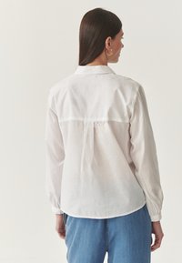Camicia bianca a maniche lunghe con colletto, caratterizzata da un cucitura a yoke sulla schiena e orlo arrotondato, indossata con jeans blu. Tessuto fluido e leggero.