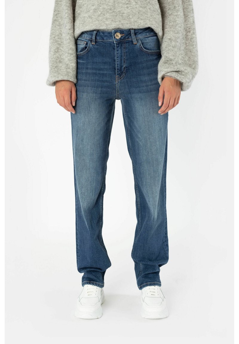 Dámské modré denimové džíny se rovnými nohavicemi, s bledými detaily, pěti kapsami a zapínáním na knoflík, v kombinaci s bílými teniskami.