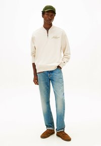 Felpa beige chiaro con cerniera e polsini a costine, abbinata a jeans blu slavati e mocassini marroni. Un berretto verde completa l'outfit.