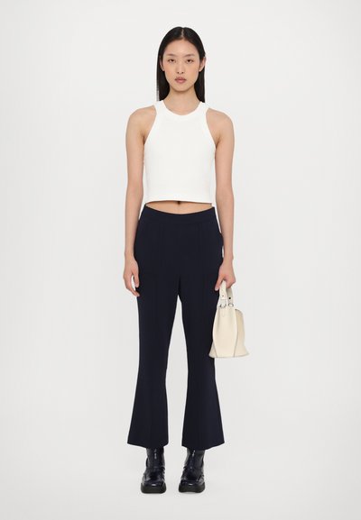 Hvid ærmeløs crop top med høj hals; mørkeblå skræddersyede flare bukser; holder en lille off-white taske; skinnende sorte ankelstøvler.