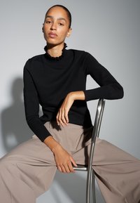 Haut noir à manches longues avec un col à volants, associé à un pantalon beige à jambes larges, assise sur une chaise argentée devant un fond gris.