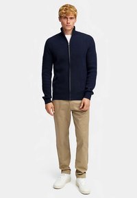 Marineblauwe geribde cardigan met ritssluiting en hoge kraag, gedragen over een wit overhemd, gecombineerd met beige broek en witte sneakers.