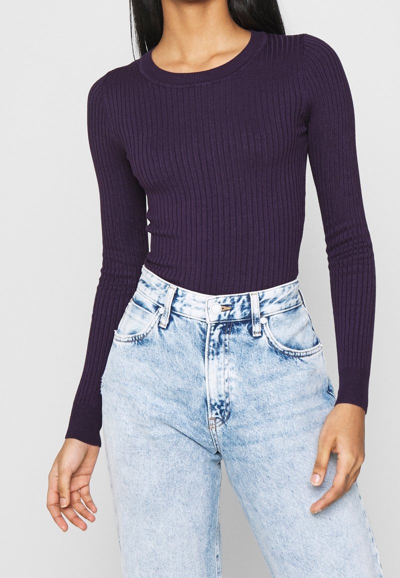 Femme portant un pull violet côtelé à manches longues et ajusté, rentré dans un jean en denim bleu clair taille haute.