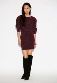 Bordeauxkleurige glinsterende jurk met pofmouwen, nauwsluitend silhouet en lengte boven de knie, gecombineerd met zwarte suède laarzen tot aan de knie.