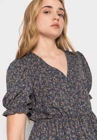 Vestido floral em tecido azul marinho com decote em V, mangas bufantes e cintura franzida. Pequenos padrões florais em vermelho e bege por toda a peça.