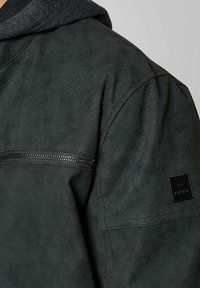 Veste en cuir noir perforé avec une capuche grise, dotée d'une poche zippée sur la poitrine et d'une étiquette en tissu texturé sur l'épaule.