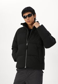 Musthave must värvitud puhkejakk kõrge kraega, lukuga ees ja horisontaalse tikandi mustriga. Omab küljetaskuid ja sileda tekstuuri.