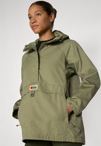Vrouw in een groene Fjällräven jas met capuchon, met handen in de voorzakken, die iets omhoog kijkt tegen een effen lichte achtergrond.