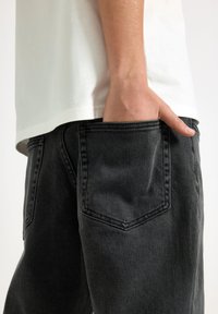 Jeans in denim nero con una texture morbida, caratterizzati da un design con tasca posteriore, indossati con una maglietta bianca. I jeans hanno un taglio dritto.