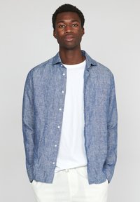 Camicia button-up blu con texture, realizzata in materiale leggero, indossata sopra una t-shirt bianca, con una vestibilità rilassata e colletto a punta.