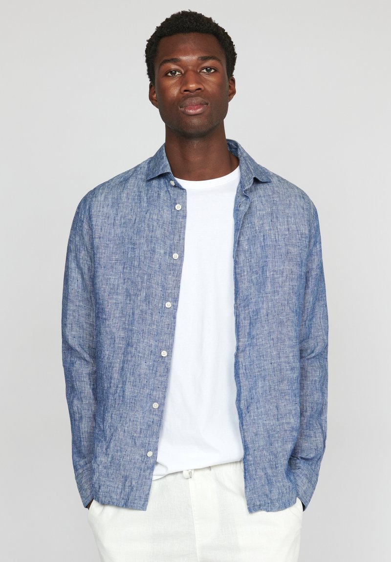 Camicia button-up blu con texture, realizzata in materiale leggero, indossata sopra una t-shirt bianca, con una vestibilità rilassata e colletto a punta.