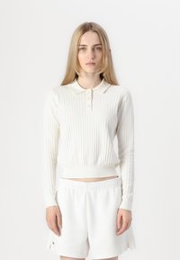Polo blanco de manga larga con canalé, cuello y tapeta con botones, combinado con pantalones cortos blancos con cinturilla elástica.