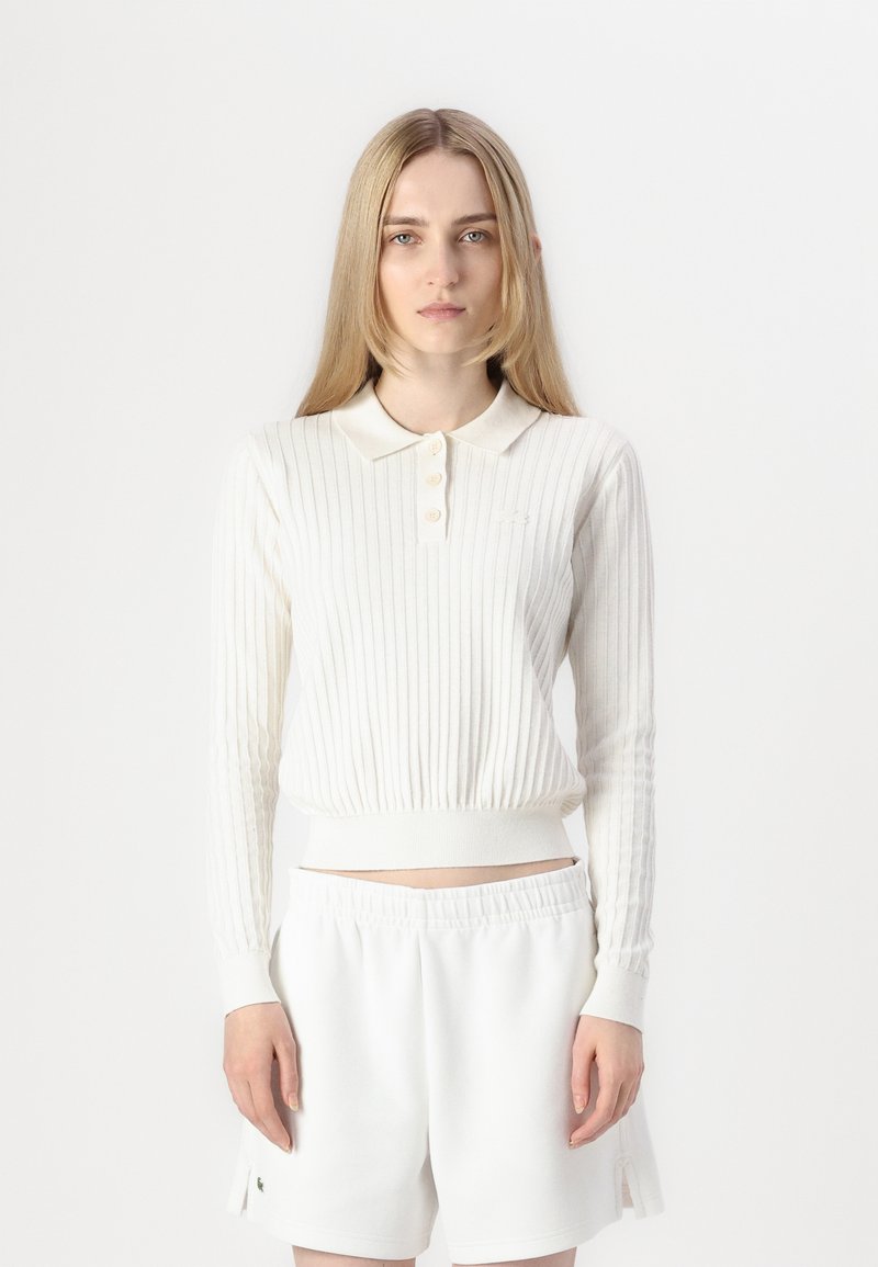 Polo blanco de manga larga con canalé, cuello y tapeta con botones, combinado con pantalones cortos blancos con cinturilla elástica.