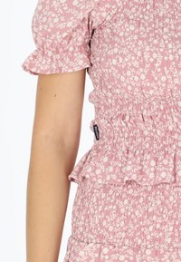 Bluse i pink med blomsterprint, der har korte pufærmer, smocket struktur og en bølget kant. Lappen viser "SEASIDE" på siden.