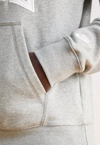 Szara bluza z kapturem wykonana z miękkiego materiału, posiadająca przednią kieszeń, ściągacze na rękawach oraz biały logo Nike na lewym rękawie.