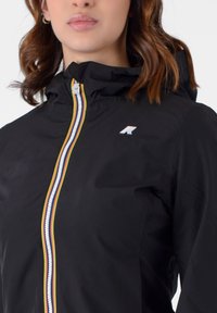 Giacca leggera nera con cappuccio, dotata di una cerniera multicolore e un piccolo logo sul petto. Texture liscia e design minimalista.