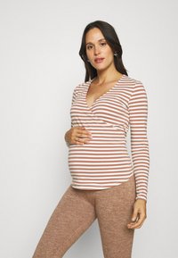 Gestreiftes, langärmliges Umstandstop in Hellpink und Beige mit Wickeldesign. Kombiniert mit braunen, strukturierten Leggings.