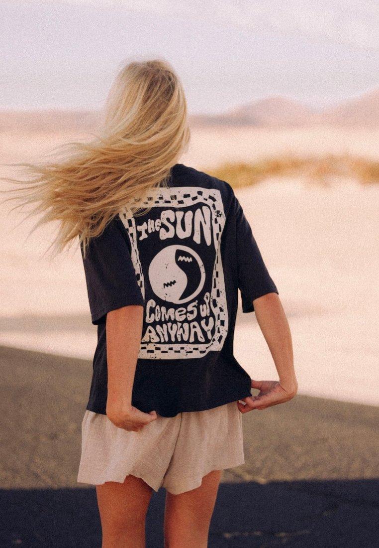 Czarna, oversize koszulka z białym nadrukiem graficznym, na którym widnieje napis "Słońce wschodzi I TAK", z okrągłym wzorem i kontrastującą, kratkowaną bordiurą.