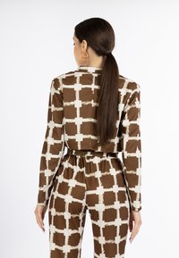 Bruine outfit met een patroon en een wit rasterontwerp, met lange mouwen, een riem op de taille en een gladde textuur. Cirkelvormige oranje oorbellen zichtbaar.