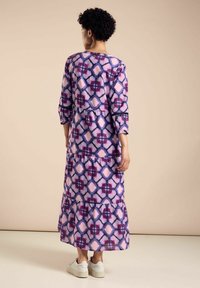 Langes lila Kleid mit geometrischem Muster in Pink und Marineblau, ausgestattet mit gestuften Rüschen und 3/4-Ärmeln. Getragen mit weißen Sneakers.