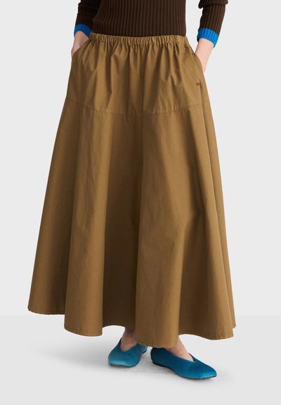 Personne portant une longue jupe marron camel avec une taille élastique et des chaussures bleues sans lacets, mains dans les poches avec un pull marron foncé et bleu.