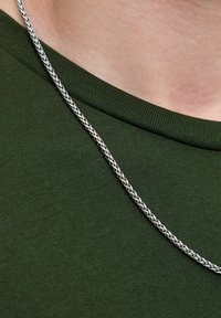 Srebrny łańcuszek na szyję ułożony na ciemnozielonym, teksturowanym materiale. Łańcuch składa się z przeplatanych ogniw, podkreślających błyszczące metaliczne wykończenie.