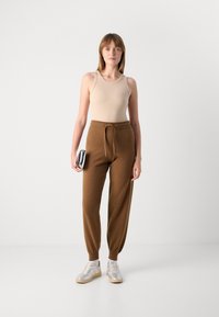 Top beige senza maniche, pantaloni jogger in maglia marrone con vita regolabile e sneaker chiare con accenti metallici; in piedi contro uno sfondo semplice.