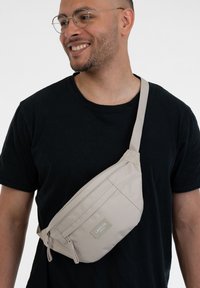 Borsa a tracolla beige con un design elegante, realizzata in tessuto con cerniere, tracolla regolabile e un piccolo etichetta del marchio sul davanti.