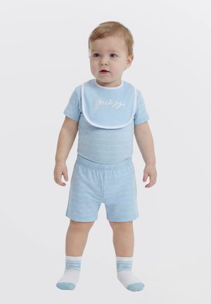 Mazs bērns, ģērbies gaiši zilā kreklā un šortos, ar saskaņotu priekšautiņu, uz kura rakstīts "guess", un baltām zeķēm ar zilām niansēm, stāv uz vienkārša fona.