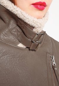 Veste en cuir marron avec un col en fausse fourrure crème, équipée de boucles en métal et d'une poche zippée. Matériau texturé, design chaud et robuste.