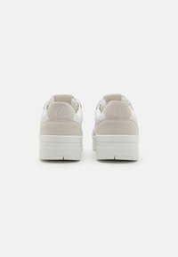 Hvit joggesko med en teksturert overdel og beige semsket hæl. Tykk hvit gummisåle og merkede hældetaljer. Minimalistisk design.