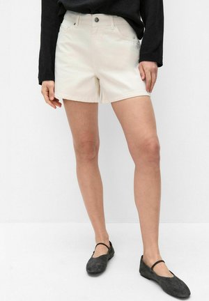 Kvinde iført off-white denimshorts, sort langærmet top og sorte Mary Jane-flade sko, stående mod en ensfarvet hvid baggrund.