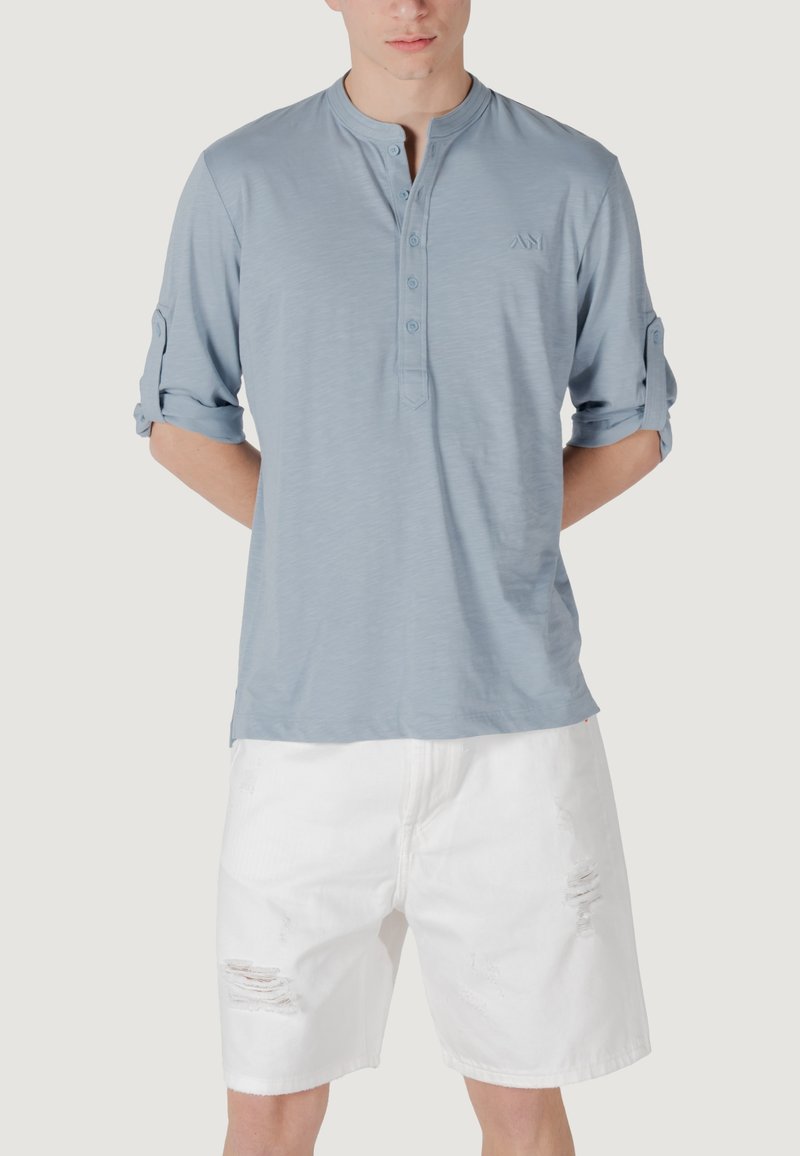 Camicetta henley a maniche lunghe di colore azzurro chiaro con patta a tre bottoni e maniche arrotolate. Indossata con pantaloni corti bianchi strappati. Design minimalista.