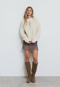 Chaqueta bomber de punto color marfil con cierre de cremallera, combinada con un vestido mini de estampado púrpura y botas de ante marrón hasta la rodilla.