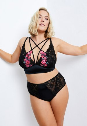 Vrouw in zwarte kanten lingerie met roze bloemrijk borduurwerk poseert tegen een witte muur met wijd gespreide armen.