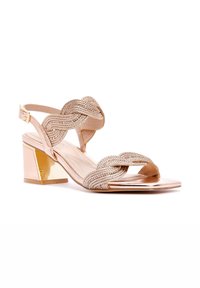 Sandal con tacco in rosa antico con cinghie intrecciate a incrocio, un tacco a blocco metallizzato e una finitura liscia e lucida. Dettaglio della fibbia regolabile alla caviglia.