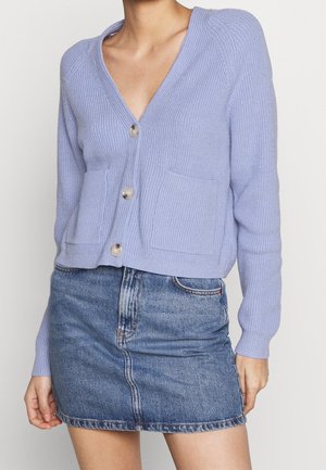 Femme portant un cardigan boutonné violet clair avec des poches avant et une mini-jupe en jean bleu.