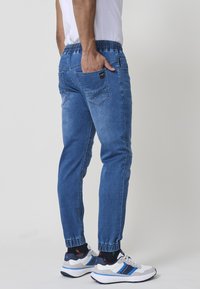 Pantalones de jogger de mezclilla azul con cintura y puños elásticos, que cuentan con bolsillos laterales y un ajuste relajado, combinados con zapatillas blancas y marinas.